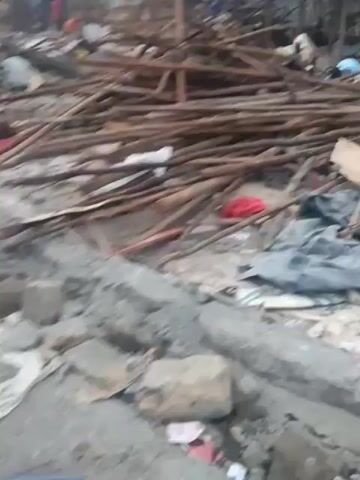 The video shows a scene of destruction, possibly after a natural disaster or demolition. Debris including wood, bricks, and pieces of metal and fabric are scattered across the ground. A partially standing structure in the background reveals a frame but missing walls. People are visible amidst the wreckage, possibly searching or assessing the damage. The environment appears urban or semi-urban, with buildings visible in the background. The overall impression is one of significant damage and disarray.