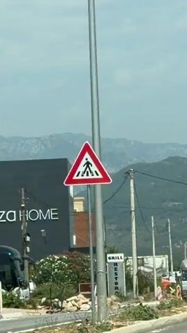 This is a vertical, medium-shot video showing a street scene with a building and an airplane in the background. In the foreground, a red and white triangular pedestrian crossing sign is visible on a metal pole. Behind it, a large dark grey building with "HOME" written in white letters is on the left. To the right of this building, there is a smaller building with brown and white accents. Further to the right, a tall vertical sign reads "GRILL RESTOR" with what appears to be a "U" or "M" missing from the end of "RESTOR." There are also power lines crisscrossing the frame and telephone poles. In the distance, mountains are visible under a cloudy sky, and an airplane is flying low and appears to be descending. Construction debris is scattered on the ground in the mid-ground, and a truck is partially visible on the right side of the frame. The overall impression is a semi-urban or roadside environment with some development and infrastructure.
