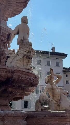 This media file captures a dynamic view of a fountain with multiple sculptures, set against a backdrop of historic buildings under a clear blue sky. The primary subjects are stone statues of human figures and mythical creatures, intricately carved and weathered by time. In the foreground, a large, ornate fountain structure is visible, with water cascading from various points, creating a sparkling, lively effect.

One prominent sculpture depicts a muscular, male figure, possibly a sea god or mythological hero, with one arm raised as if in action or greeting, while water spouts from his mouth. Another sculpture, higher up on the fountain, shows a child-like figure, turned away from the camera, also surrounded by water. The elaborate stonework of the fountain itself, with its detailed carvings and multiple tiers, suggests it is a significant historical monument.

Behind the fountain, several European-style buildings rise, characterized by their pastel-colored facades, numerous windows with shutters, and tiled roofs. The architecture suggests an urban setting in a European city, consistent with the provided location context of Trento, Italy. The bright sunlight indicates it is daytime, likely mid-morning or afternoon, with fair weather and a few scattered clouds visible in the distance. The overall mood conveyed is one of vibrant public life and artistic heritage, with the water adding a sense of energy and refreshment to the scene. No visible text is present in the media file.