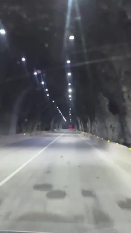 The video shows a view from inside a car driving through a tunnel. The tunnel has rough rock walls and a ceiling lined with lights. The road ahead is paved and well-lit, with a white lane marker. In the distance, there are red lights visible, possibly indicating traffic or construction. A white car passes by on the left lane. The camera is positioned at dashboard level, suggesting it's mounted on the car's interior. The footage is slightly shaky, consistent with a moving vehicle.
