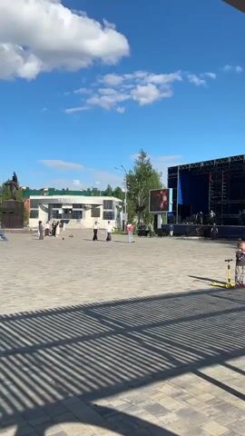 The video shows a town square on a sunny day with a blue sky and scattered clouds. In the foreground, shadows from an overhead structure create a striped pattern on the paved ground. A young child stands near a yellow scooter on the right side of the frame, facing toward the center of the square. Several people are milling about in the middle ground, some walking and others standing in small groups. In the background, a large stage is set up, covered in blue fabric with people moving on and around it. A large screen displays an image. Buildings are visible in the distance.