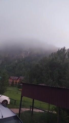 A misty mountain looms over a dense forest. A few houses with red roofs are nestled among the trees. A dark brown awning stretches across the foreground. A white car is parked to the left. The sky is overcast and appears to be dawn or dusk.
