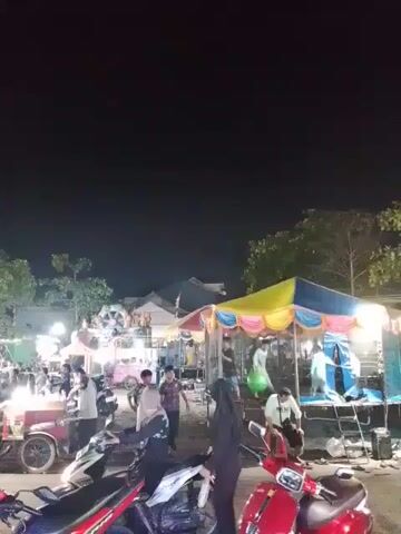The video shows an outdoor night market scene.  Several food vendor carts are visible, as are carnival-style rides or games.  People of various ages are milling around, some wearing casual clothes and others in traditional attire.  Scooters are parked near the camera. The lighting is bright, with strings of lights and spotlights illuminating the stalls. Trees and buildings can be seen in the background.
