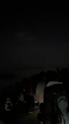 The video shows a group of people standing in what appears to be a dark, outdoor setting at night. The sky is visible above, with some hints of clouds. The foreground features a person with a backpack, facing away from the camera and towards the crowd. Some people are holding up their phones, presumably taking photos or videos of something in the distance. The lighting is very dim, making details difficult to discern. There are silhouettes of trees and other figures scattered throughout the scene.