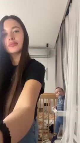 A woman with long brown hair, wearing a black t-shirt and jeans, stands in a room and makes gestures towards the camera. Behind her, a baby stands in a white crib, holding onto the side and looking at the camera. The room has a neutral color scheme, with sheer white curtains hanging on the right and an air conditioning unit mounted on the wall. A black pendant light hangs from the ceiling. Music is playing in the background.