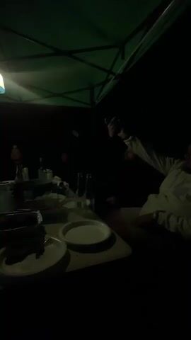 A group of men are gathered around a table in a dark, outdoor setting, possibly a campsite. A green canopy tent provides overhead cover, illuminated by a hanging light fixture. On the table are plates, food in serving dishes, bottles, and glasses, suggesting a meal or gathering. To the left, a projection screen displays a football match, with one man facing the screen and appearing engaged. The overall atmosphere appears relaxed and social.