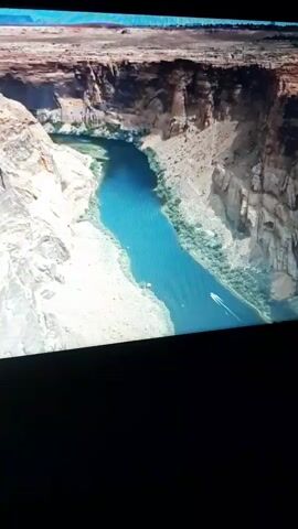 An aerial view shows a boat traversing a wide, blue river that flows between towering, rocky canyon walls. The canyon walls are a mix of light and dark brown rock, with patches of green vegetation clinging to the lower slopes. The river has a smooth surface, reflecting the bright blue sky. The boat is small and white, leaving a wake behind it as it moves downstream. The scene is vast and natural, emphasizing the scale of the canyon and the river.