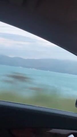 The video is filmed from inside a car, looking out the passenger window. The landscape outside consists of a large body of water with a distant shoreline featuring mountains. The sky is overcast with a pale blue and grey hue, suggesting either early morning or late evening. The water is a vibrant turquoise color, and there are a few distant lights visible on the shore. The interior of the car is partially visible, including a dark-colored dashboard and a side mirror. The car appears to be in motion, as indicated by the slight blur in the footage and the passing of what looks like a power line tower. The song playing in the background is a slow-paced ballad.
