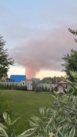 A plume of pink-tinged smoke billows into the sky. In the foreground, a grassy yard is visible, with a small shed to the left and a fence behind it. A blue roof of a building is partially visible to the left of the shed. Trees are visible in the background, and the sky is overcast with clouds.