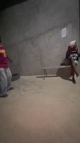 Inside a dimly lit room with unfinished concrete walls and a dirt floor, three people are present. One person sits on a small metal bench against the back wall. They are wearing a basketball jersey labeled "DEL MAR 20" and have a head covering on. Another individual, wearing a maroon basketball jersey with the number 23, dances in the open space. A third person wearing a white shirt and patterned shorts walks past the dancer. A tire is leaning against the wall. A piece of paper is stuck to the wall. A cell phone and other objects are on the bench.
