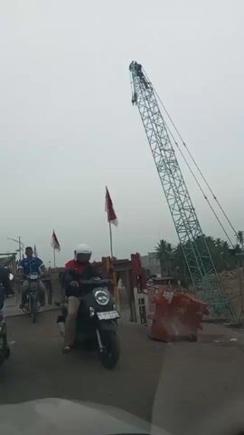 The video shows a traffic scene on a bridge under construction. Several motorcycles and a car are visible, moving slowly. A large green crane looms in the background, and red and white flags, possibly national flags, are displayed along the bridge. The sky is overcast. The camera appears to be inside a vehicle.