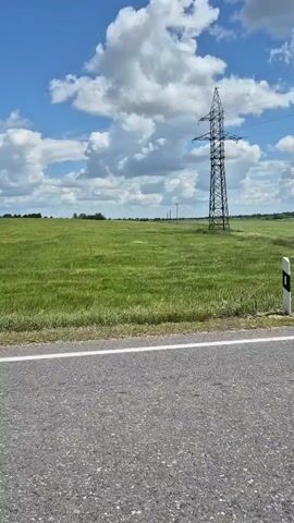 A static shot shows a rural landscape under a partly cloudy blue sky. In the foreground, a paved road runs horizontally across the bottom of the frame, with a white line marking the edge. Beyond the road is a vast, flat green field. A tall, latticed metal power line tower stands prominently in the middle ground of the field, to the right of center. A white and black road marker is visible on the right edge of the frame, next to the road. The overall impression is one of open space and a clear day.