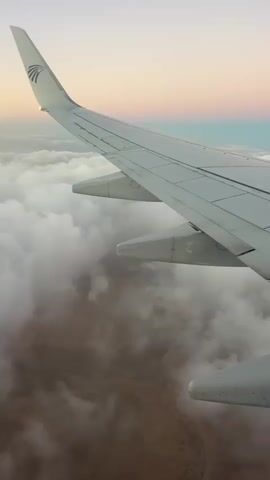 The video shows a view from an airplane window. The wing of the airplane is in the foreground, with a desert landscape visible through the clouds below. The sky is a gradient of pink, orange, and blue, suggesting either sunrise or sunset. The airplane's logo is visible on the tail fin. The clouds are thick and cover most of the ground. The video is short and appears to be a still image with slight movement.