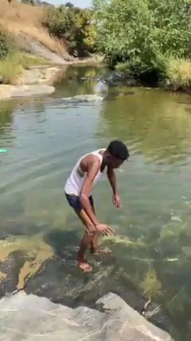 A young man with dark hair, wearing a white tank top and dark shorts, stands in a shallow, clear river. He is bent over, reaching into the water with both hands. He then rises, holding a translucent white object, possibly fabric. The river flows over smooth, grey rocks. The surrounding environment features green trees and a grassy hillside. The scene is captured under bright sunlight.