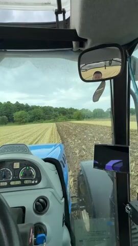 The video shows the interior of a tractor cab, with the view looking out through the windshield and side mirror. The tractor is plowing a field, with freshly turned earth visible. In the side mirror, a person's face is reflected, presumably the driver. The landscape outside is rural, with fields and trees under a cloudy sky. There are various instruments and controls visible inside the cab, including a steering wheel, dashboard gauges, and a tablet mounted on the right side. A hanging air freshener is also visible. The video is filmed from a first-person perspective, giving the impression of being in the driver's seat of the tractor.