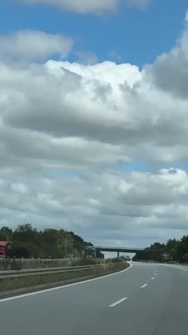 The video shows a POV shot from a moving vehicle on a highway. The road ahead curves to the right. On the left side of the road, there is a grassy embankment with trees and bushes. A guardrail separates the embankment from the highway. Over the embankment, a concrete overpass spans the highway. A large truck is visible on the highway, to the left of the overpass. The sky is cloudy with patches of blue.