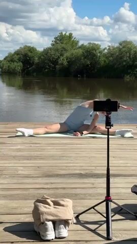 A young woman performs a split on a wooden dock by a river. She is wearing a light blue athletic outfit and white socks. A smartphone is mounted on a tripod in front of her, recording her workout. Her arms are extended upwards in one pose, then she transitions to placing her hands on the dock in front of her. In the foreground, a pair of white sneakers and a beige garment are placed on the dock. The background features a river, lush green trees, and a cloudy sky.