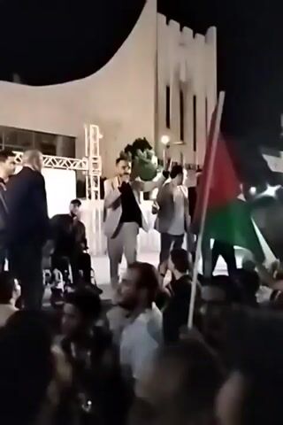The video shows a crowd of people gathered at night, likely for a celebration or rally. In the foreground, a person waves a Palestinian flag.  In the center of the video, a man in a light gray suit stands on a small stage, gesturing and addressing the crowd. Behind him, other people are on the stage.  The crowd appears to be mostly men, with some younger individuals visible.  The setting is an outdoor area with buildings and streetlights in the background. The lighting is a mix of artificial light from the stage and streetlights.