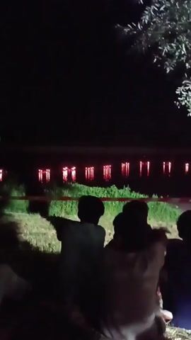 A nighttime video shows several people sitting on the ground facing a body of water. In the distance, a building with many illuminated windows is visible across the water. Red and white striped tape is stretched across the foreground, presumably marking a boundary. The overall atmosphere is dark, with the light from the building and a nearby tree providing the primary illumination.
