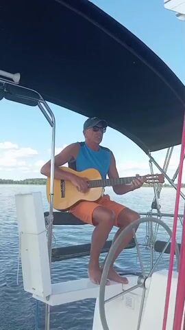 A man is playing a guitar on a sailboat. He is wearing a blue sleeveless shirt, orange shorts, and a baseball cap. He is also wearing sunglasses. The sailboat is on the water, and the sky is blue with some clouds. The sun appears to be shining, suggesting it is daytime. The man looks relaxed and happy as he plays his guitar. There is a steering wheel in the foreground of the image, indicating the boat is in motion or about to be. The background shows a shoreline with trees.