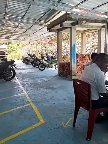 The video shows an outdoor area with a covered roof. In the foreground, a person in a white shirt is seated in a red plastic chair. To the left, another person is seated in a similar chair. Further back, two other people are seated in chairs. There are motorcycles parked in the background. A pile of bricks is visible near a concrete structure. The ground is paved and has yellow markings. The environment appears to be somewhat rustic with a stone wall in the background.