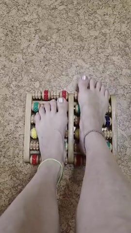 A first-person perspective video shows a pair of feet on a wooden foot roller massager. The feet are positioned on top of the roller, with the toes pointing upwards and the heels slightly elevated. The roller massager is made of wood and has several small wooden rollers that are colored red, green, and yellow. The feet have painted toenails and are adorned with anklets. The background appears to be a textured cork floor. The video shows subtle movement of the feet on the roller, suggesting a massage action.
