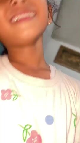 This is a close-up, shaky video of two young girls. The girl in the foreground, who appears to be younger, is looking upwards and laughing. She has dark hair pulled back, small gold hoop earrings, and is wearing a white t-shirt with colorful floral and animal prints. Her neck and face are shiny, possibly from sweat. Her head is tilted back, revealing her open mouth and teeth as she laughs.

The second girl, who appears slightly older, is positioned behind the first. She has dark hair and is wearing a light-colored top. She has her arms around the first girl's shoulders, and her face is partially obscured as she smiles and possibly laughs along.

In the background, there are some visual cues that help establish the location and setting. A teal-colored wall is visible, along with what looks like a framed picture or artwork on another wall. There are also some blurred-out objects that could be furniture or bedding, suggesting an indoor setting. The lighting appears to be natural, and the overall mood is joyful and playful. The camera movement and focus suggest this was captured in a candid moment, likely at home. The timestamp suggests the video was captured in the early morning or late evening, but the actual time of day cannot be definitively determined without more context. There is no visible text in the video.
