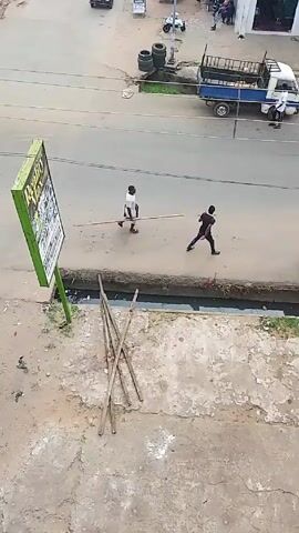 A high-angle shot shows two individuals walking across a street. One, wearing white, carries a long pole. The other, in black, walks behind him. In the background, there are parked vehicles, pedestrians, and a small pickup truck. A green sign stands near the bottom left corner of the frame, and some wooden poles lie on the ground nearby. The surrounding environment appears to be a commercial area with buildings and parked vehicles.