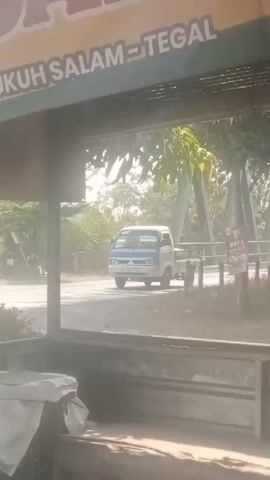 The video pans across a dusty roadside scene. A white pickup truck with blue accents drives by on a paved road. In the foreground, there are wooden structures, possibly part of a roadside stall or warung, with signs that are partially visible. One sign clearly reads "WARUNG Bu ISA". Trees and foliage are visible in the background, and the overall lighting suggests it is daytime, possibly late morning or afternoon, with a hazy or dusty atmosphere.