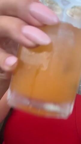 This is a short, vertical video clip. The subject is a young woman with dark hair styled in two braids. She is wearing a red tank top and a dark baseball cap. Her skin appears to be lightly tanned, and she is making a kissing face at the camera. In her left hand, which is held up to the camera, she is holding a clear glass filled with an orange-colored liquid, possibly juice. Her fingernails are painted a pale pink.

The background suggests an indoor setting with a large window or glass door, through which a blurred view of the outdoors can be seen. The lighting indicates it is daytime. The woman is seated, and the perspective is a close-up selfie. The overall mood appears casual and playful. There is no visible text in the video. Given the provided location context of Byurakan, Armenia, this video may have been taken at a hotel, restaurant, or private residence with a view.