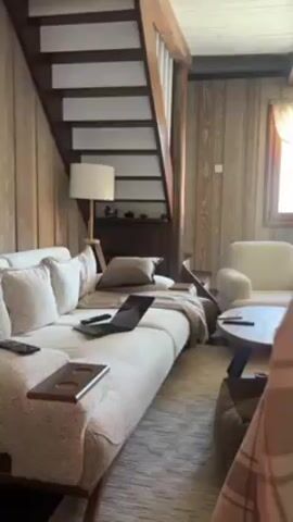 The video shows a living room with rustic decor. The walls are paneled with light-colored wood. There's a large, light-colored sofa with multiple pillows and a throw blanket. A laptop, phone, and remote control are on the sofa. A round coffee table sits in front of the sofa, and there's a staircase in the background. The video pans to show a fireplace with a fire burning inside, along with decorative items on the mantelpiece, including a clock, a wooden box, and a lamp.