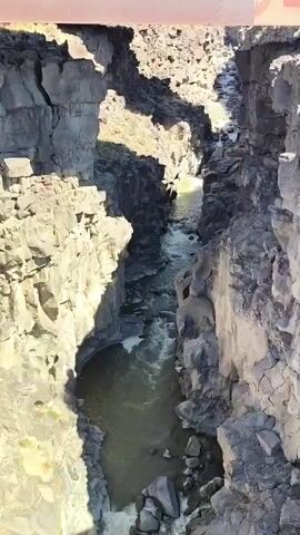 A high-angle shot looks down into a narrow gorge where a river flows. The gorge walls are made of dark, rough-hewn rock, with sharp, angular formations. Sunlight casts shadows on the canyon walls, highlighting their textured surfaces and deep crevices. The water in the river is a murky green, with white foam indicating it is moving rapidly and encountering obstacles. The river narrows as it moves further into the gorge. The top edge of the frame shows a glimpse of what appears to be a bridge structure, made of dark metal. The overall scene suggests a rugged, natural landscape carved by the persistent flow of water.
