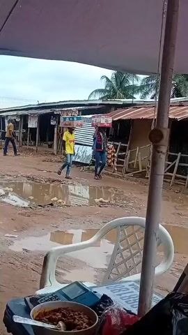 The media file depicts an outdoor scene in Yelwa, Nigeria, likely a market or roadside area. The ground is muddy and waterlogged, suggesting recent rainfall or poor drainage. In the background, several small structures with corrugated metal roofs line the path, resembling market stalls or shops.

Two individuals are walking from left to right across the muddy ground. The person on the left is wearing a yellow t-shirt and blue jeans, carrying a rectangular object on their head. The person on the right is wearing a blue jacket over a patterned top, blue jeans, and also carrying something on their head. Both appear to be walking with purpose. Another person in a yellow shirt is visible further in the background on the left.

In the foreground, a white plastic chair is partially visible. On a surface in front of the chair, there is a bowl of food, a calculator, and some other items, suggesting a vendor or a resting spot. A large umbrella, likely providing shade, dominates the top of the frame. A portion of an A-frame sign with the text "LIZZY BLISS COMMUNICATION" is also visible in the foreground. Text on the umbrella appears to be "PalmPay". The lighting suggests it is daytime, possibly overcast given the lack of strong shadows. The overall impression is of a functional, bustling, and somewhat weathered environment typical of a local market in Nigeria.