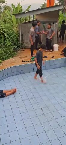 The video shows a group of young people at a swimming pool. In the foreground, a boy is lying on his back in the shallow end of the pool, kicking his legs. Another boy is in the middle of the pool, swimming on his stomach. In the background, several other young people are standing or sitting around the edge of the pool. Some of them are talking and laughing. One boy is leaning against a wall, and another is sitting on the edge of the pool with his feet in the water. The pool area appears to be outdoors, with vegetation visible in the background. The pool itself has blue tiled walls and a tiled floor. There is a concrete area around the pool, with some debris and scattered flip-flops. A shelter with a corrugated metal roof is visible to the left. The overall impression is of a casual, recreational setting.