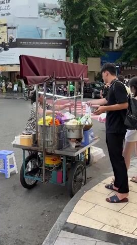 A street vendor operates a food cart on a sidewalk. The cart is equipped with various containers and cooking apparatus, including steaming pots filled with food items. The vendor, an adult female, is actively preparing food. A young male customer is interacting with the vendor, receiving an item. Another young female customer stands nearby, holding a cup. The environment is an urban street with a pedestrian crossing in the background, suggesting a commercial or busy area. Traffic, including motorbikes and cars, is visible in the background, along with buildings and advertising displays. The overall scene depicts a typical street food transaction in an outdoor setting.