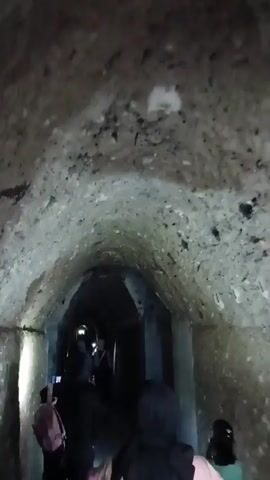 A group of people are walking through a narrow, dimly lit tunnel with a rough, stone ceiling. Several individuals are wearing headlamps or holding flashlights, illuminating the path ahead and the textured walls. Some of the people are wearing headscarves, suggesting a cultural context. The tunnel appears to be an ancient or historical structure, possibly underground, with arched ceilings and a worn, uneven floor. The camera is moving with the group, giving a first-person perspective of their exploration.
