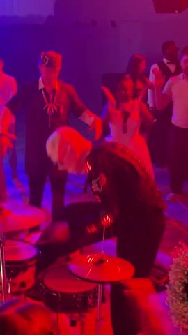 The video shows a drummer wearing a white mask and a black costume with glowing red accents, playing drums on a stage. The drummer is leaning forward and appears to be performing energetically. Behind the drummer, several people are visible, some dancing and others watching. The environment is dimly lit, with red and purple stage lighting creating a festive atmosphere. The person on the left is wearing a black outfit with a spider emblem.