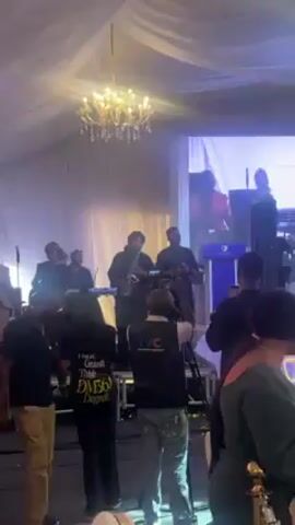 A band performs live music at an event in a tented venue. The band includes a saxophone player in the center, a singer on the right, and several other musicians. A woman in a pink jacket speaks on a screen behind the band. Attendees stand and watch, some filming with their phones. A TVC News camera operator records the performance. A chandelier hangs above the band.