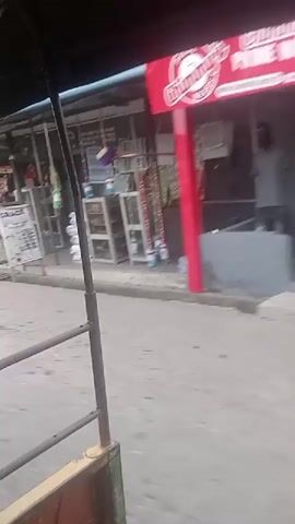 A vehicle with an open side is driving down a paved street. A collection of small businesses and houses line the street. People are walking on the sidewalk, and one is seen being dragged along the ground. A gray car is parked on the side of the road. There are stacks of green crates in front of a gate. A sign reading "FOOD IS READY" is visible. The video appears to be taken in an urban environment.