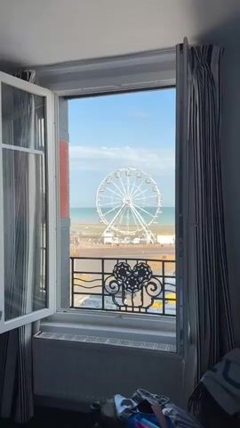 The video opens from an indoor perspective looking out through an open window. The window frame is white with some dark detailing and there are striped curtains visible on either side. The view outside is a bright, sunny day with a clear blue sky. Dominating the mid-ground is a large Ferris wheel, white and circular, with many cabins attached. Beyond the Ferris wheel is the ocean, appearing calm and blue, with a faint line of beach visible in front of it. In the foreground, below the Ferris wheel, is a parking lot filled with various cars. A black decorative metal railing is visible just in front of the window, adorned with a circular, ornate pattern. The architecture of a building, possibly a hotel or apartment, is visible on the right side of the frame. A person is walking on the sidewalk in the far right of the frame.