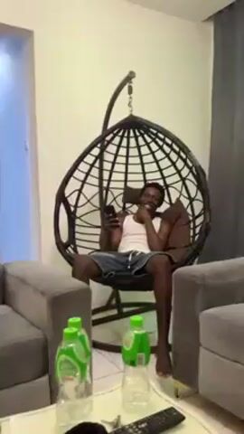 A man with dark skin and short hair sits in a hanging chair in a living room. He is wearing a white tank top and blue shorts, and he is holding a smartphone. There are two gray sofas on either side of the chair, and a white coffee table in front of them. On the table are three bottles of sparkling water and a black remote control. The walls are white, and there is a dark gray curtain in the background. The man is smiling and gesturing with his hands.