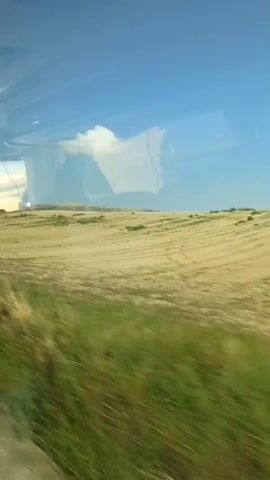 This video shows a landscape viewed through a window, likely from a moving vehicle. The scene consists of a vast, dry, harvested field with visible rows of tilled earth. In the distance, a large array of solar panels is set up on a grassy hillside under a bright blue sky with a few scattered clouds. The foreground is blurred, indicating motion, and shows some dry grasses or reeds. Reflections are visible on the window.