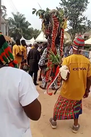 The video shows a cultural performance in what appears to be a village setting. The central figure is a person in a tall, elaborate costume with red and white patterns, topped with leaves and what looks like a small animal carcass. They're surrounded by people; some are wearing yellow shirts with "CCYA" on the back, and one wears a colorful striped hat. Others are dressed in everyday clothing. The performers are walking down a dirt road, and there's a tent and other people in the background. The event seems to be taking place outdoors on a cloudy day.