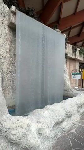 The video shows a slow-motion close-up of a water feature in what appears to be an indoor environment. The water falls in a translucent sheet from a horizontal bar at the top of a large, flat, rectangular panel. The panel has faint vertical lines, possibly from the material it's made of or the way the water flows. The water collects in a shallow basin at the bottom, which is surrounded by a rough, stone-like structure. The background includes a wooden ceiling with beams and some greenery, suggesting an interior space like a mall or atrium. A sign with text and a logo is visible on the right side, further indicating a commercial or public setting. The water flow is smooth and continuous, creating a gentle cascading effect. The lighting is soft, and the overall impression is one of tranquility.