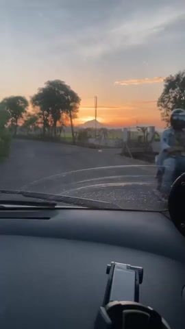The video shows a view from inside a vehicle, looking out the windshield at a rural landscape during sunrise. The sun is visible on the horizon, casting an orange glow across the sky. Trees line a paved road, and there are fields and a fence in the background. A black object, possibly a phone mount, is visible on the dashboard in the foreground. The video is short, lasting only a few seconds, and appears to be filmed while the vehicle is in motion.