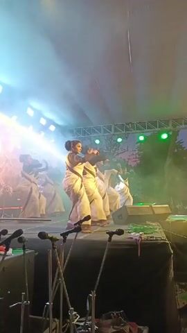 The video shows a group of women performing a dance on a stage. They are all wearing white saris and have their hair pulled back. The stage is lit with bright lights, and there is some smoke or fog in the air. In the foreground, there are several microphones on stands. The dancers are in various poses, some with their arms raised and others with their hands clasped. The overall impression is of a coordinated and graceful performance.