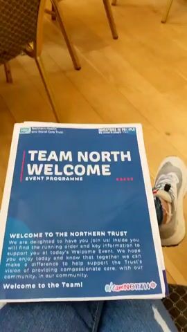 The video shows a room with a wooden floor and chairs arranged in rows. In the foreground, a program booklet titled "TEAM NORTH WELCOME EVENT PROGRAMME" is visible. The booklet is blue and white and has text that reads "WELCOME TO THE NORTHERN TRUST". In the background, a group of people are seated, facing a screen and a presenter. The presenter appears to be a man in a white shirt. The room has a high, sloped ceiling and a mural depicting a cityscape on the wall. The overall scene suggests a welcome event or conference.