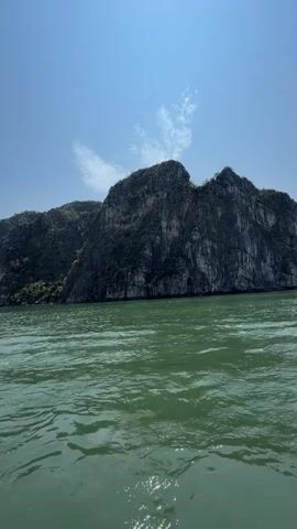 The video captures a scenic daytime view of Tuan Chau Ward, Vietnam, likely from a moving boat. The foreground features choppy, greenish water, indicating movement. In the midground and background, majestic limestone karsts rise dramatically from the water, their dark, rugged surfaces partially covered with lush green vegetation. The sky above is a clear, bright blue with a few wispy white clouds. The overall impression is one of a beautiful, tranquil natural landscape under a sunny sky. No people or visible text are present in the video.