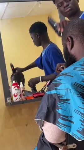 A barber, wearing a blue shirt, is cutting a client's hair in a salon. The client, wearing a blue and black patterned cape, is seated in a chair facing away from the camera. The barber is reflected in a mirror that is mounted on a yellow wall. The mirror shows the barber using clippers to cut the client's hair. There are several bottles of hair products on a shelf below the mirror.