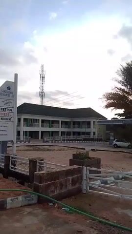 The video shows a gas station and a building. On the left is a tall sign that reads "SAKE GLOBAL RESOURCES LTD." Below that, it lists "PETROL," "DIESEL," and "KEROSENE." The gas station has pumps visible. Behind the gas station is a two-story building with green trim. A cell tower is visible in the background. The sky is cloudy.